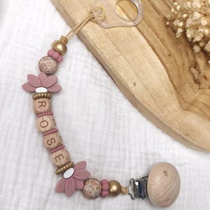 Op de afbeelding: Een speenkoord met een reeks kralen in de kleuren oudroze, goud en naturel hout. De kralen bevatten twee siliconen bloemvormen, ronde gespikkelde kralen en vier houten blokjes die de naam ROSE spellen. Een doorzichtige plastic lus en een houten clip zijn aan de uiteinden van het koord bevestigd.