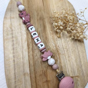 Peut inclure: Une attache-sucette composée de perles en silicone dans des tons vieux rose et blanc, incluant une perle en forme de faon, une perle en forme de fleur et des perles lettres blanches formant le prénom LENA. L'attache se termine par un fermoir en métal.