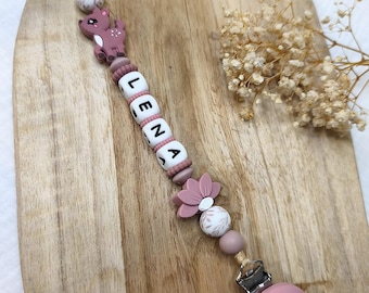 May include: A pacifier clip featuring a series of silicone beads in shades of dusty rose and white, including a deer-shaped bead, a flower-shaped bead, and white letter beads spelling the name LENA. The clip ends with a round, embossed metal fastener.