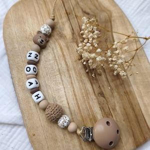 Peut inclure: Une attache-tétine personnalisée pour bébé avec une perle en forme d'ours brun, des perles en bois et des perles de lettres épelant un nom. La tétine est marron et le clip est en métal. Sur une planche en bois.
