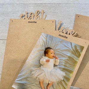 K&ouml;nnte beinhalten: Ein kleines M&auml;dchen in einem wei&szlig;en Tutu und einem wei&szlig;en Strampler liegt auf einer hellgrauen Decke. Das Baby schaut nach rechts im Bild. Das Bild ist mit einem braunen Kartonrahmen umrahmt. Die Worte "acht Monate" stehen oben am Rand und die Worte "zw&ouml;lf Monate" stehen unten am Rand.