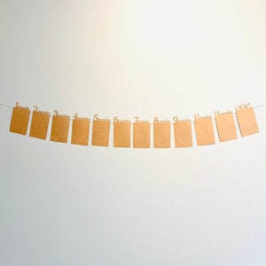 K&ouml;nnte beinhalten: Ein braunes Papierbanner mit den Zahlen 1 bis 12, jeweils mit dem Wort "Monate" darunter. Das Banner ist an einer wei&szlig;en Wand befestigt.