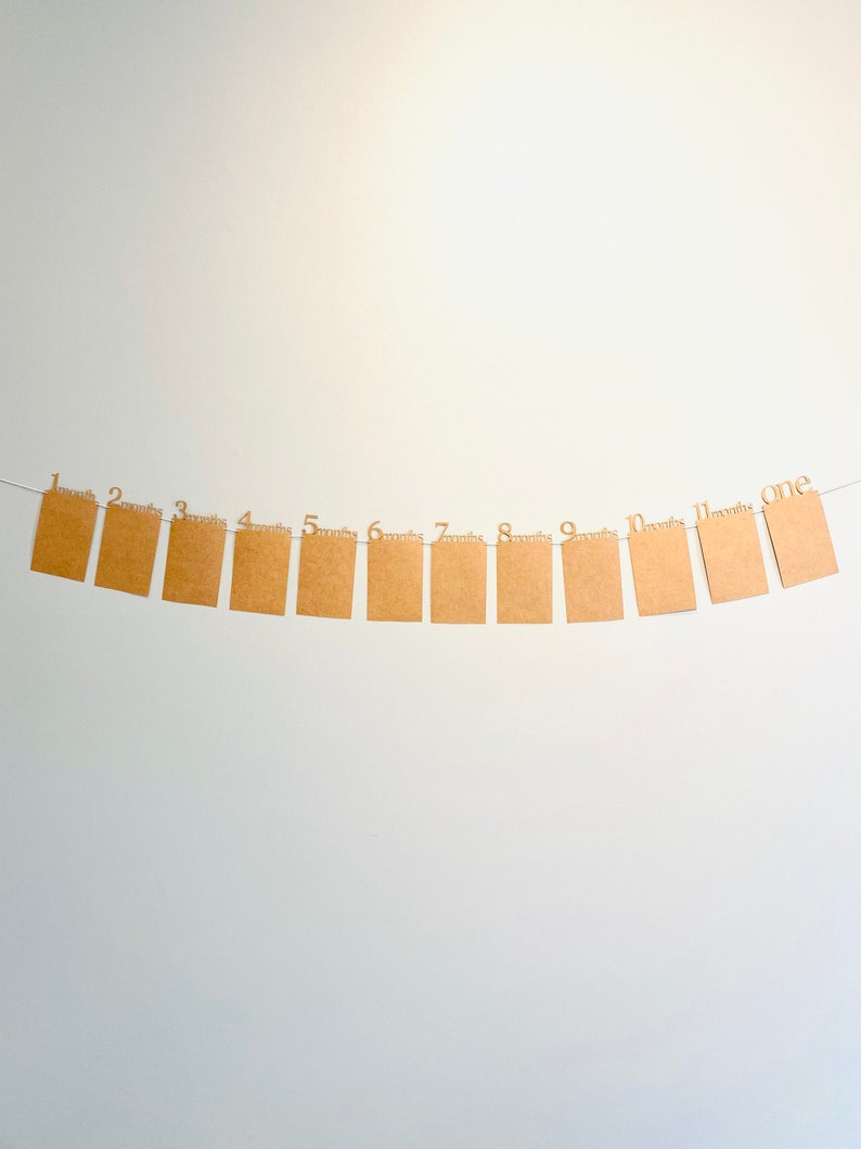 K&ouml;nnte beinhalten: Ein braunes Papierbanner mit den Zahlen 1 bis 12, jeweils mit dem Wort "Monate" darunter. Das Banner ist an einer wei&szlig;en Wand befestigt.