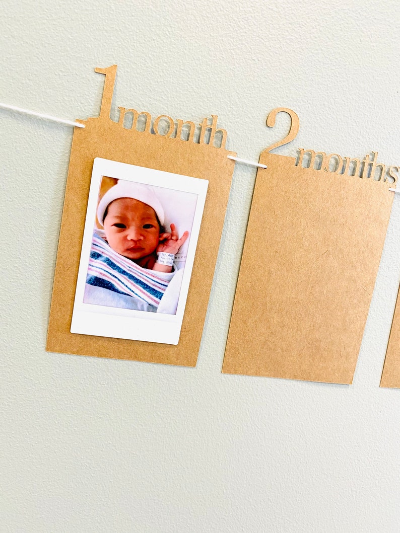 K&ouml;nnte beinhalten: Ein braunes Papierbanner mit den Worten "1 Monat" und "2 Monate" darauf gedruckt. Das Banner hat eine wei&szlig;e Schnur daran befestigt und ein Foto eines Babys in einem gestreiften Strampler befindet sich im ersten Rahmen.