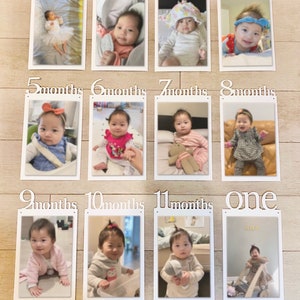 K&ouml;nnte beinhalten: Eine wei&szlig;e, h&ouml;lzerne Fotoanzeige f&uuml;r monatliche Meilensteine mit 12 Rahmen, jeder mit einem Foto eines Babys in verschiedenen Altersstufen. Die Rahmen sind mit dem Monat des Babyalters beschriftet, von 1 Monat bis 12 Monate. Der letzte Rahmen ist mit "eins" beschriftet.