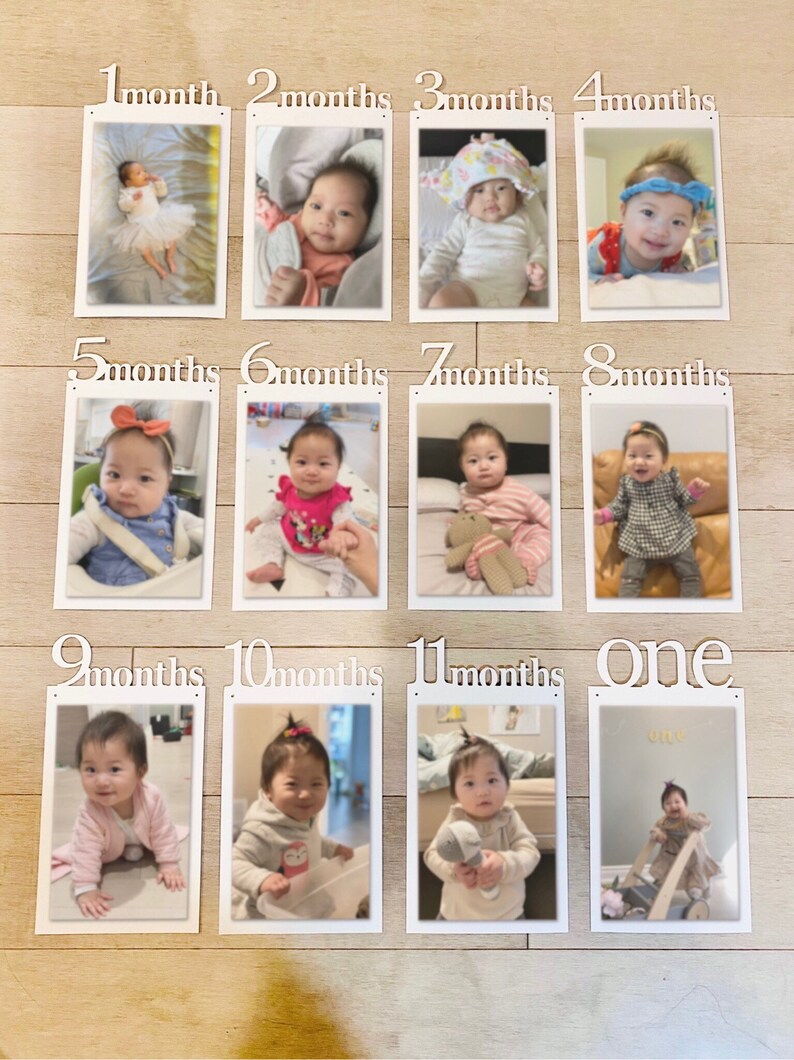 K&ouml;nnte beinhalten: Eine wei&szlig;e, h&ouml;lzerne Fotoanzeige f&uuml;r monatliche Meilensteine mit 12 Rahmen, jeder mit einem Foto eines Babys in verschiedenen Altersstufen. Die Rahmen sind mit dem Monat des Babyalters beschriftet, von 1 Monat bis 12 Monate. Der letzte Rahmen ist mit "eins" beschriftet.