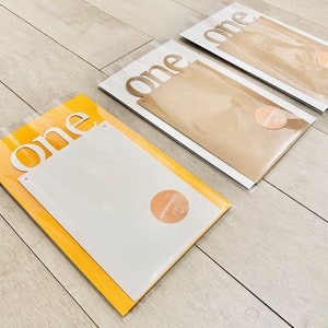 K&ouml;nnte beinhalten: Drei wei&szlig;e Papp-Ausschnitte des Wortes "one" in einer modernen Schriftart. Die Ausschnitte sind in durchsichtigen Plastikt&uuml;ten mit braunem Papierr&uuml;cken verpackt. Jede T&uuml;te hat einen kleinen runden Aufkleber mit dem Wort "celebration" darauf.