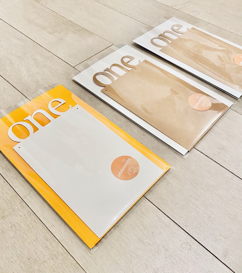 K&ouml;nnte beinhalten: Drei wei&szlig;e Papp-Ausschnitte des Wortes "one" in einer modernen Schriftart. Die Ausschnitte sind in durchsichtigen Plastikt&uuml;ten mit braunem Papierr&uuml;cken verpackt. Jede T&uuml;te hat einen kleinen runden Aufkleber mit dem Wort "celebration" darauf.
