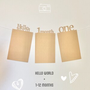 May include: A brown paper banner with the words "Hello World", "1 month", and "one" printed on it. The banner is strung up with white string and there are two white hearts and a white camera drawn on the background.