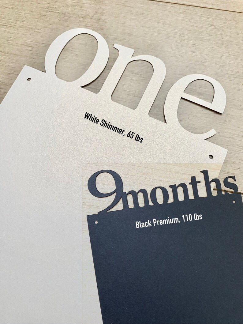 K&ouml;nnte beinhalten: Zwei Holzschildchen mit den ausgeschnittenen W&ouml;rtern "one" und "9 months". Das Schildchen "one" ist aus wei&szlig;em Schimmerkarton und das Schildchen "9 months" aus schwarzem Premiumkarton.