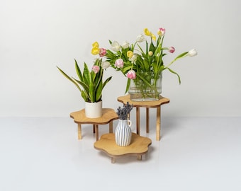 May include: Three wooden plant stands in the shape of a cloud, with a white potted plant, a bouquet of pink and yellow tulips in a clear glass vase, and a small blue and white striped vase with lavender flowers.