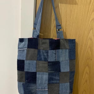 May include: A patchwork denim tote bag with a variety of blue and gray squares. The bag has long denim straps and gold-colored metal hardware. The bag is hanging on a wooden door.