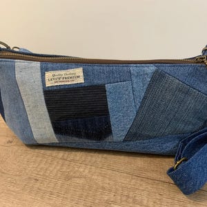 May include: A denim crossbody bag with a patchwork design. The bag features various shades of blue denim, a brown zipper, and a matching adjustable strap. A small label reads "Quality Clothing LEVI'S PREMIUM".