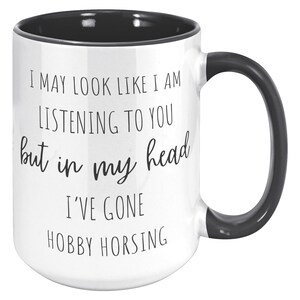 May include: White ceramic coffee mug with a black rim and handle. The mug has black text that reads "I may look like I am listening to you but in my head I've gone hobby horsing."