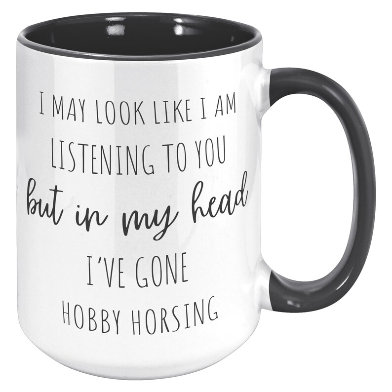 May include: White ceramic coffee mug with a black rim and handle. The mug has black text that reads "I may look like I am listening to you but in my head I've gone hobby horsing."