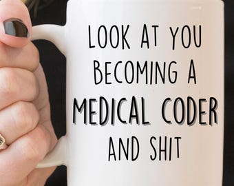 May include: White ceramic mug with the text "LOOK AT YOU BECOMING A MEDICAL CODER AND SHIT" in black, bold letters. The mug has a curved handle and is held by a person.
