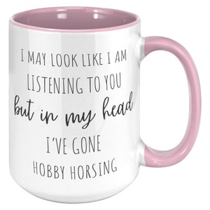 May include: White ceramic coffee mug with a pink rim and handle. The mug has black text that reads "I may look like I am listening to you but in my head I've gone hobby horsing."