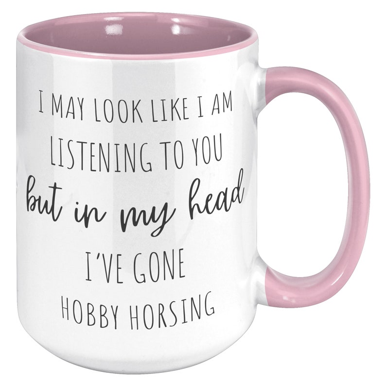 May include: White ceramic coffee mug with a pink rim and handle. The mug has black text that reads "I may look like I am listening to you but in my head I've gone hobby horsing."