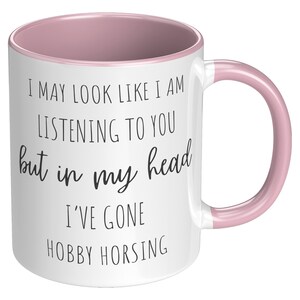 May include: White ceramic mug with a pink rim and handle. The mug has black text that reads "I may look like I am listening to you but in my head I've gone hobby horsing."