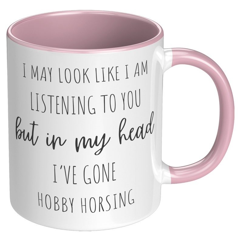 May include: White ceramic mug with a pink rim and handle. The mug has black text that reads "I may look like I am listening to you but in my head I've gone hobby horsing."
