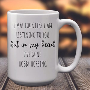 May include: White ceramic coffee mug with black text that reads "I may look like I am listening to you but in my head I've gone hobby horsing."