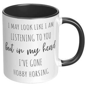 May include: White ceramic coffee mug with a black rim and handle. The mug has black text that reads "I may look like I am listening to you but in my head I've gone hobby horsing."