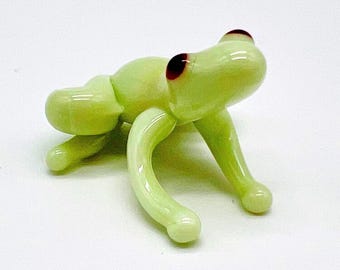 May include: A light green glass grasshopper figurine with red eyes. The insect has curved legs and a rounded body. The figurine is on a white background.
