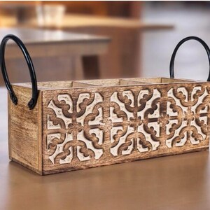 May include: A wooden utensil holder with a carved, cream-colored geometric design. The holder has three compartments and two black metal handles. The wood has a natural, light brown finish, and the design is repeated across the front.