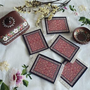May include: Set of five red and gold patterned coasters with white fringe. The coasters are on a white tablecloth with a floral design. The coasters are made to look like miniature rugs.