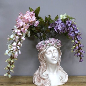 May include: A white ceramic bust of a woman with long flowing hair, used as a planter. The bust is decorated with a pink floral wreath and has purple and pink flowers cascading from the top.