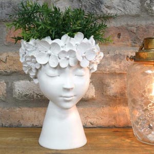 May include: A white ceramic head-shaped planter with closed eyes, decorated with a floral crown. The planter contains green foliage. The design is set against a brick wall and wooden surface, creating a decorative and artistic display.