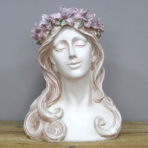 May include: White plaster bust of a woman with long, flowing hair. She is wearing a crown of pink flowers.