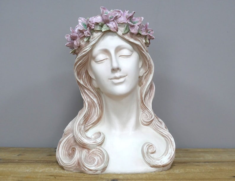 May include: White plaster bust of a woman with long, flowing hair. She is wearing a crown of pink flowers.