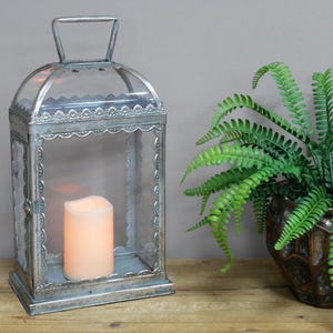 May include: A silver metal lantern with a glass pane and a decorative border. The lantern has a handle on top and a candle inside.