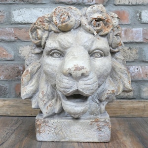 May include: A distressed stone lion head sculpture, adorned with a floral crown. The lion's face is detailed, with an open mouth and flowing mane. The sculpture, in shades of beige and brown, sits on a rectangular base, showing signs of age.