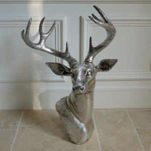 May include: A silver-coloured deer head sculpture with large antlers. The sculpture is mounted on a wall and has a detailed, textured surface. The deer head is a decorative item, suitable for home decor.