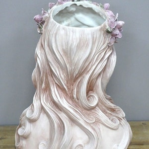 May include: A white ceramic planter shaped like a woman's bust with long, flowing hair. The planter has a floral crown with pink flowers.