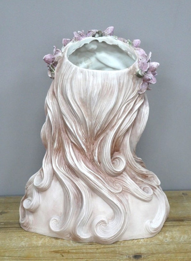 May include: A white ceramic planter shaped like a woman's bust with long, flowing hair. The planter has a floral crown with pink flowers.