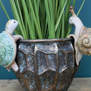 May include: A decorative planter with a textured brown finish. A green turtle and a snail with floral designs are climbing the sides of the planter.