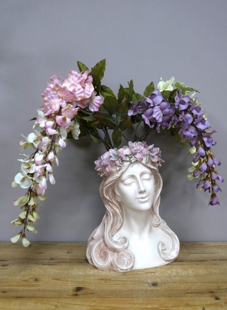 May include: A white ceramic bust of a woman with long flowing hair, used as a planter. The bust is decorated with a pink floral wreath and has purple and pink flowers cascading from the top.