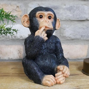 May include: A black and brown figurine of a chimpanzee with its finger to its lips, as if to say 'shhh'. The figurine is sitting on a wooden surface.