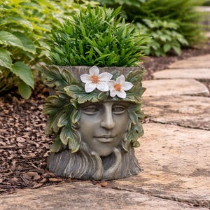 May include: A grey resin planter, sculpted as a woman's face, decorated with leaves and two white flowers. The planter holds green plants and is placed on a stone path, suitable for garden decoration.