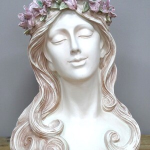 May include: White ceramic bust of a woman with closed eyes and a flower crown. The crown is made of pink and green flowers. The woman's hair is long and flowing, with a light brown color.