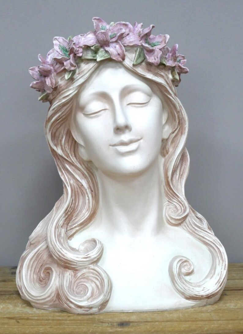May include: White ceramic bust of a woman with closed eyes and a flower crown. The crown is made of pink and green flowers. The woman's hair is long and flowing, with a light brown color.