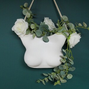 May include: White ceramic planter shaped like a bust, hanging from a white cord. The planter is filled with white roses and green eucalyptus leaves.