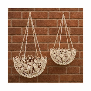 May include: Two off-white metal hanging baskets featuring intricate wirework designs of flowers and leaves. Each basket is suspended by three thin metal chains that meet at a single top hook, creating a rounded, open-frame structure for plants.