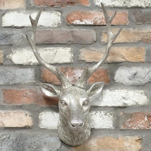 May include: Silver-coloured deer head wall decor with detailed antlers and facial features. The deer head is mounted on a multi-coloured brick wall, creating a rustic and elegant aesthetic. A decorative piece for home or cabin.