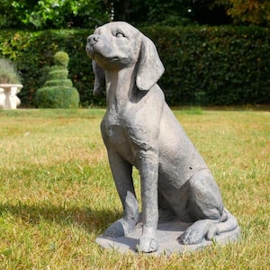 May include: A grey concrete dog statue in a seated pose, with its head raised. The statue features long, drooping ears and is positioned on a grassy lawn. A hedge and other garden elements are visible in the background.