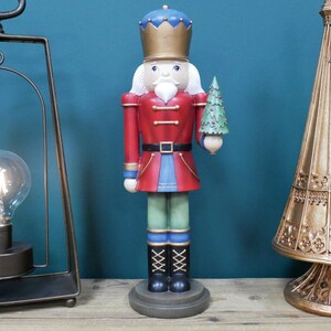 May include: A decorative wooden nutcracker figurine in a classic soldier design. The figurine is dressed in a red jacket, blue trousers, and black boots. It holds a small green Christmas tree. The nutcracker stands on a round base.