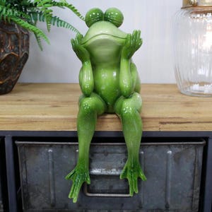 May include: A green frog figurine with a wide smile, sitting with its legs crossed and hands on its face, as if it is thinking. The frog is sitting on a wooden shelf with metal drawers.